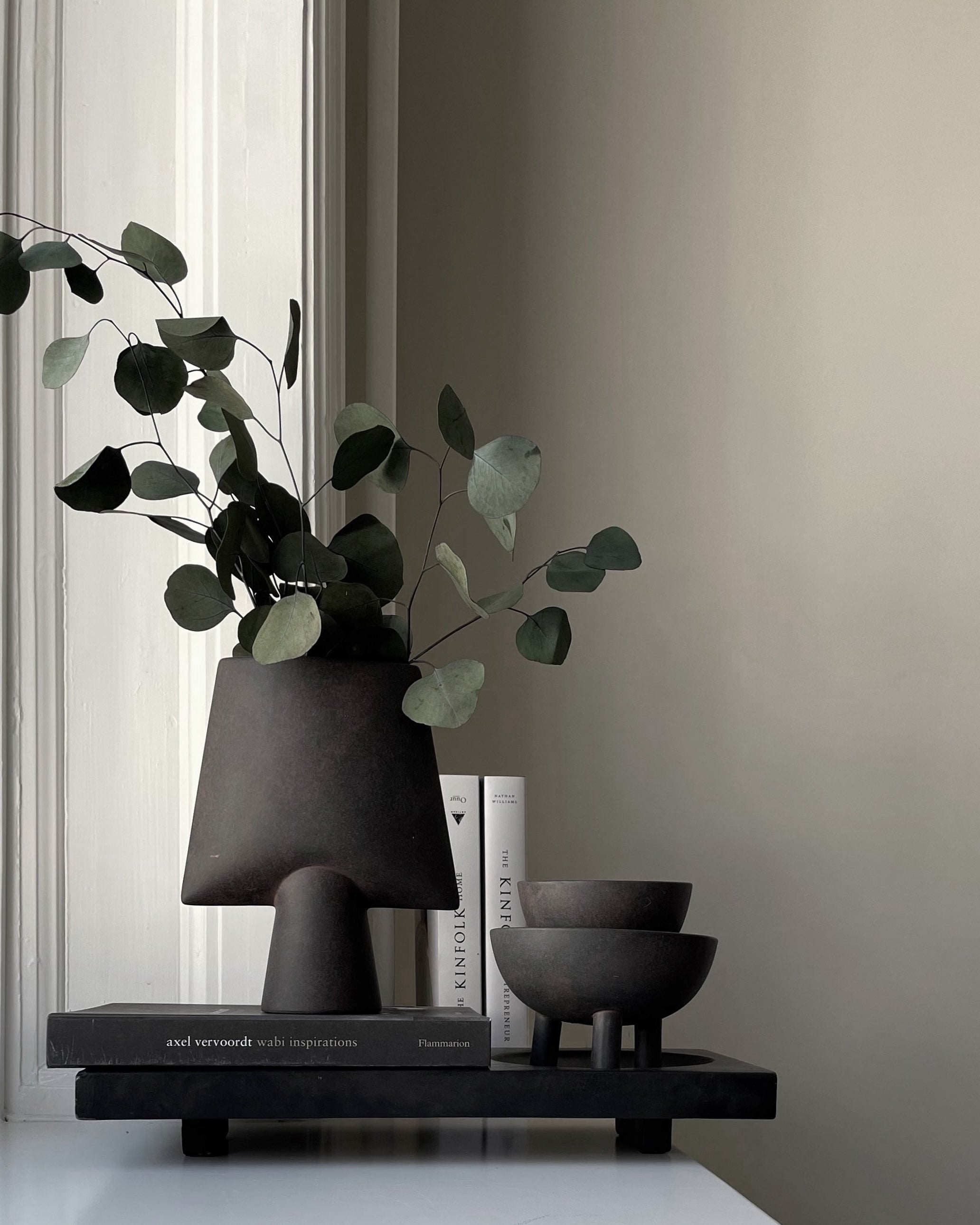 Decorative setup with black vase, books, and bowls on a shelf against a neutral wall.