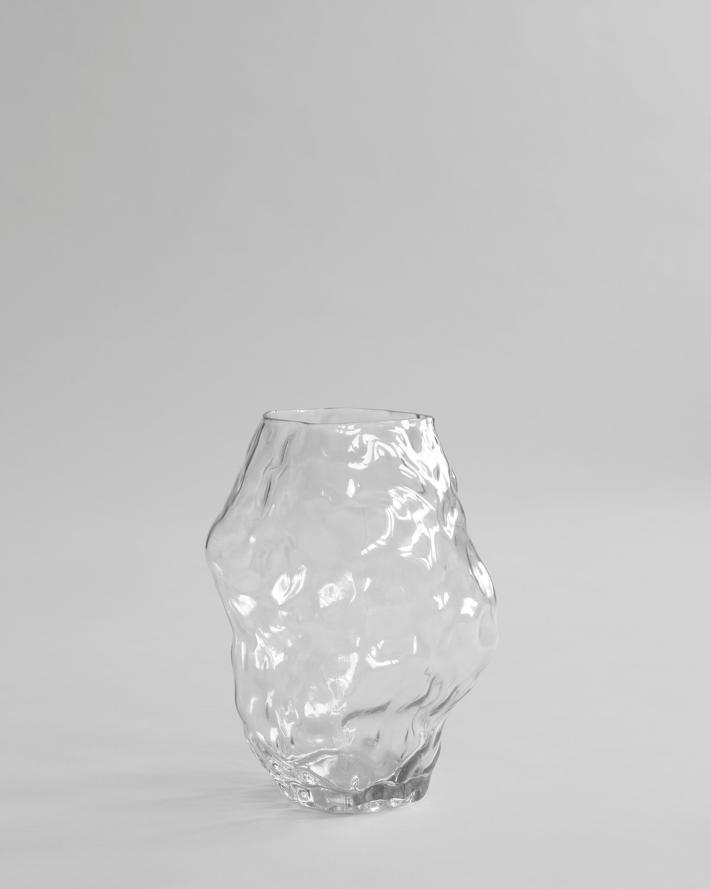 Clear glass vase on a light gray background. Katachi Vase, Medio by 101 Copenhagen. Sculptural vase inspired by Japanese “shape”, available in High Glaze Birch ceramic and Clear or Smoked Forest mouth-blown glass. Fully waterproof – curated by Numa Home.