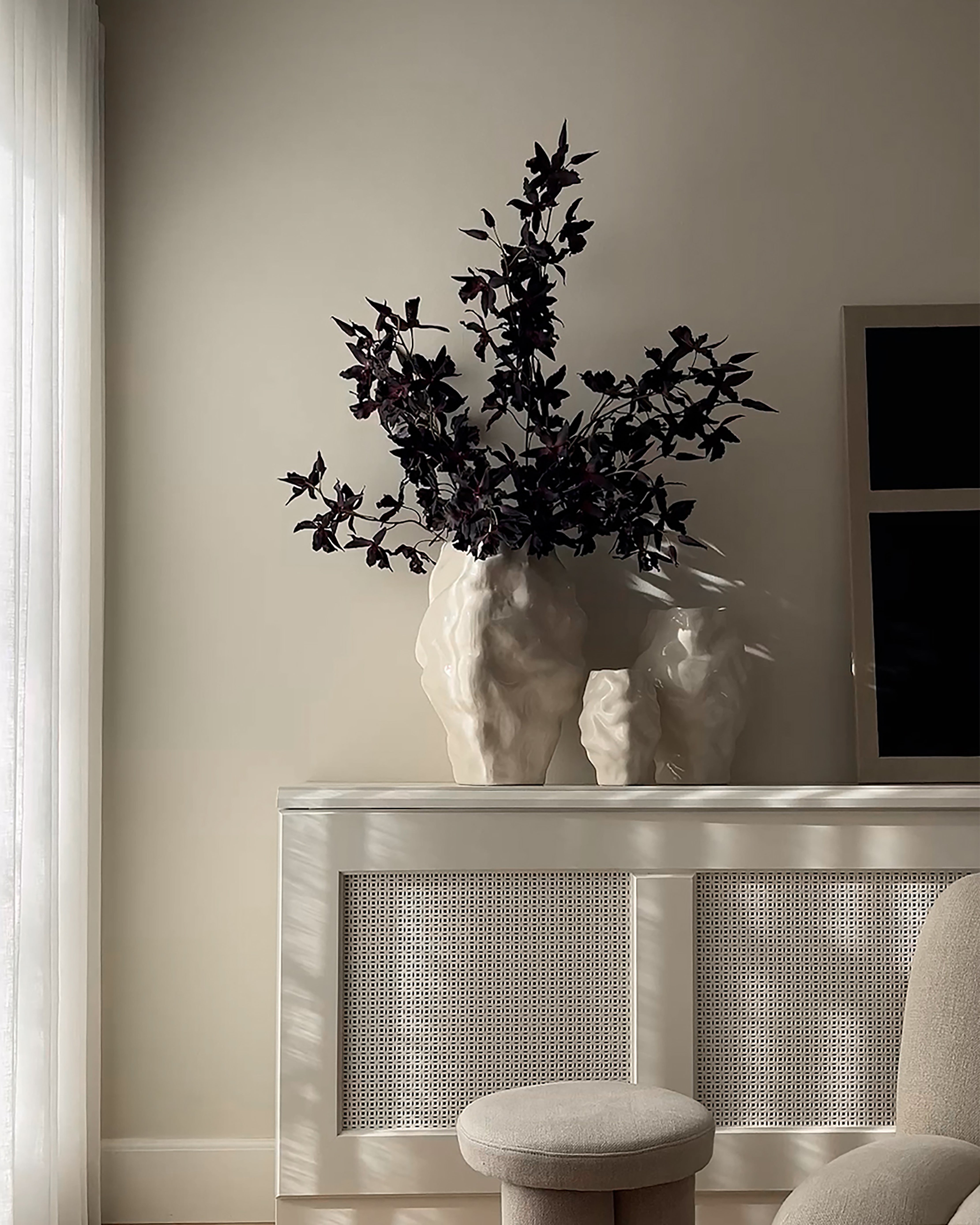 Decorative vase with dark foliage on a white console table against a beige wall. Katachi Vase, Big by 101 Copenhagen. Sculptural vase inspired by Japanese “shape”, available in High Glaze Birch ceramic and Clear or Smoked Forest mouth-blown glass. Fully waterproof, curated by Numa Home.