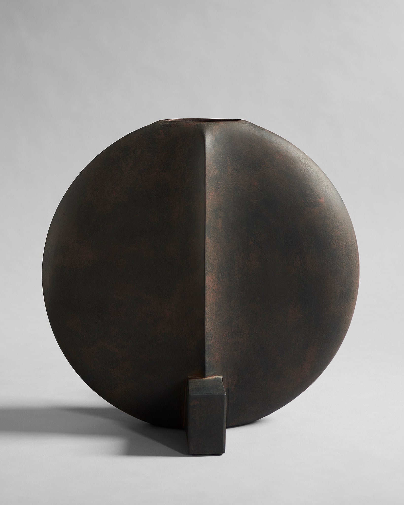 Bronze coffee color sculpture vase of a half-circle with a rectangular base on a gray background. Guggenheim Vase by 101 CPH curated by Numa Home