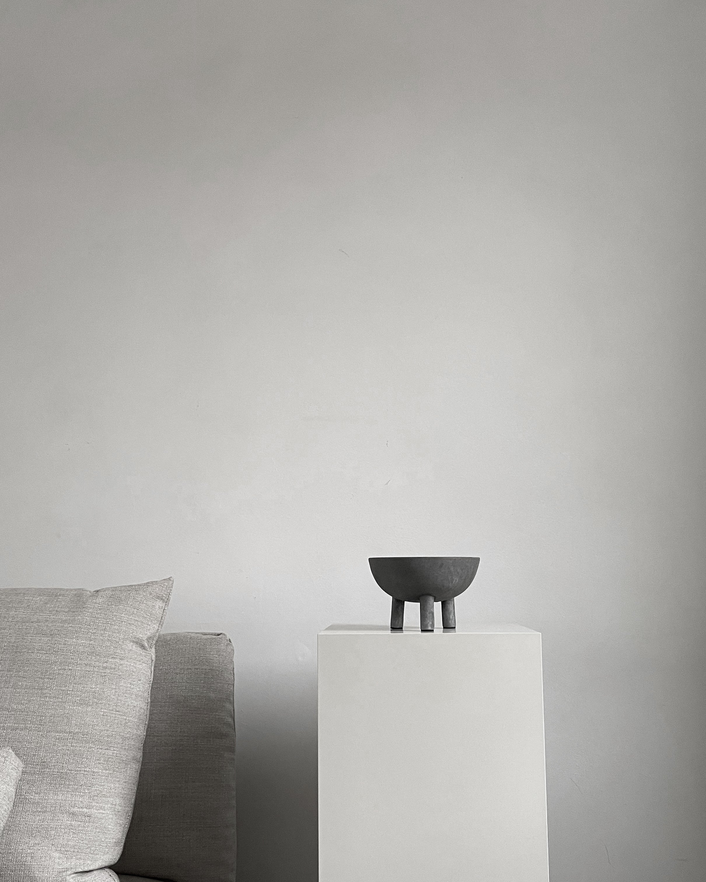 Black bowl on a white pedestal against a gray wall with a gray sofa.