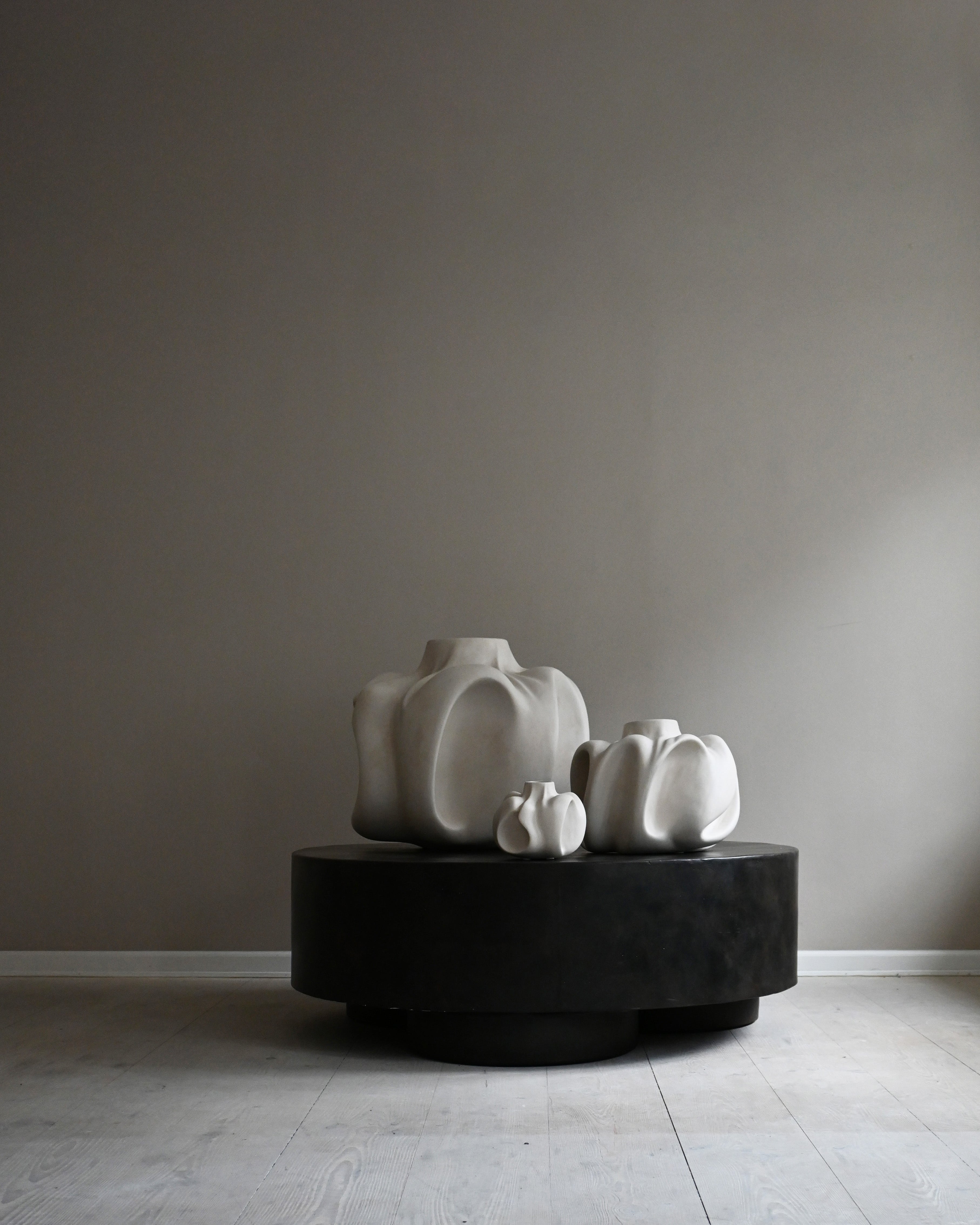 Three white ceramic sculptures on a black pedestal against a gray wall.