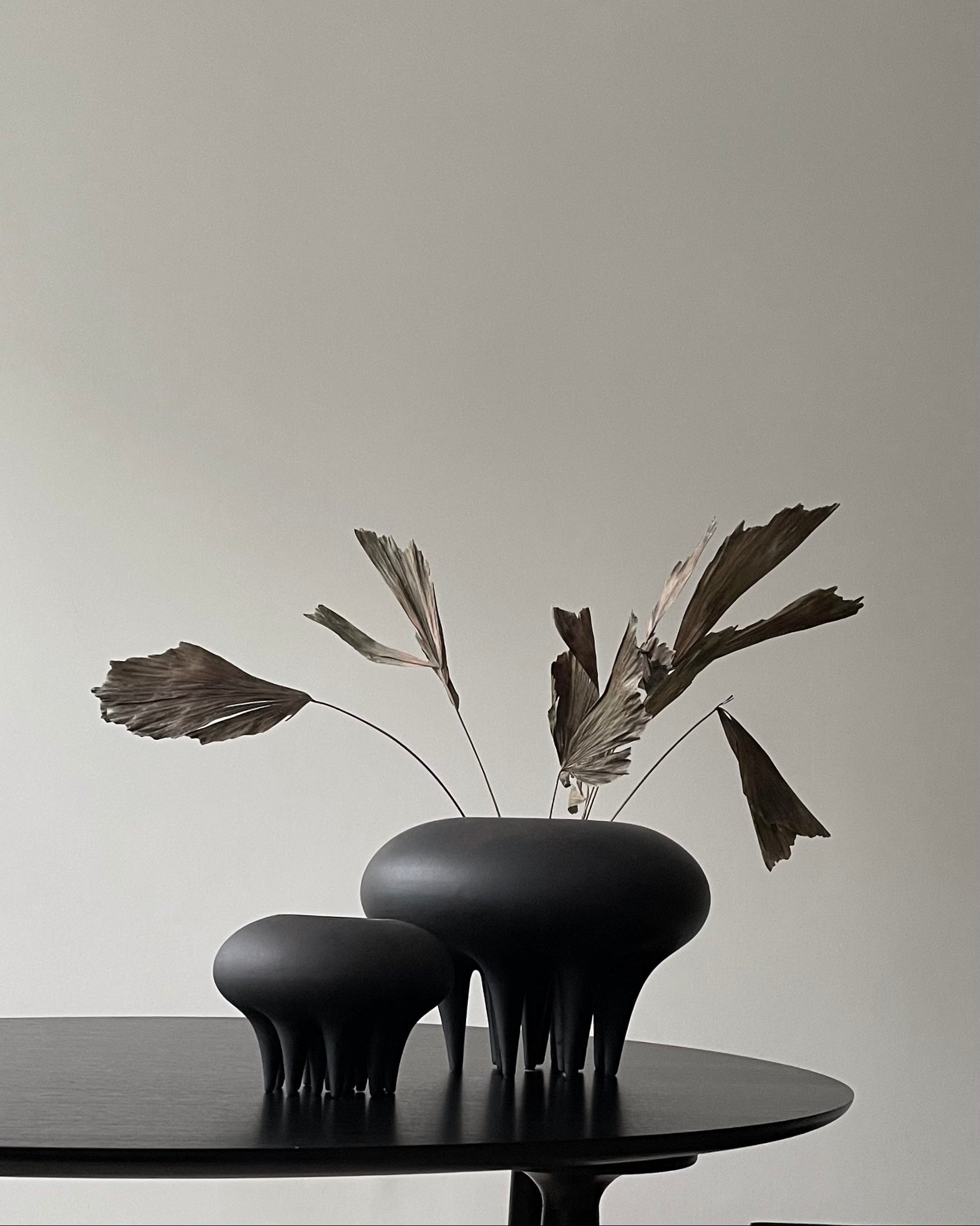 Two black vases with dried leaves on a table against a plain background. Medusa Vase, Big by 101 Copenhagen. Sculptural ceramic vase with soft oval shapes and small conical legs, hand-glazed in Coffee. Curated by Numa Home for calm, modern interiors.