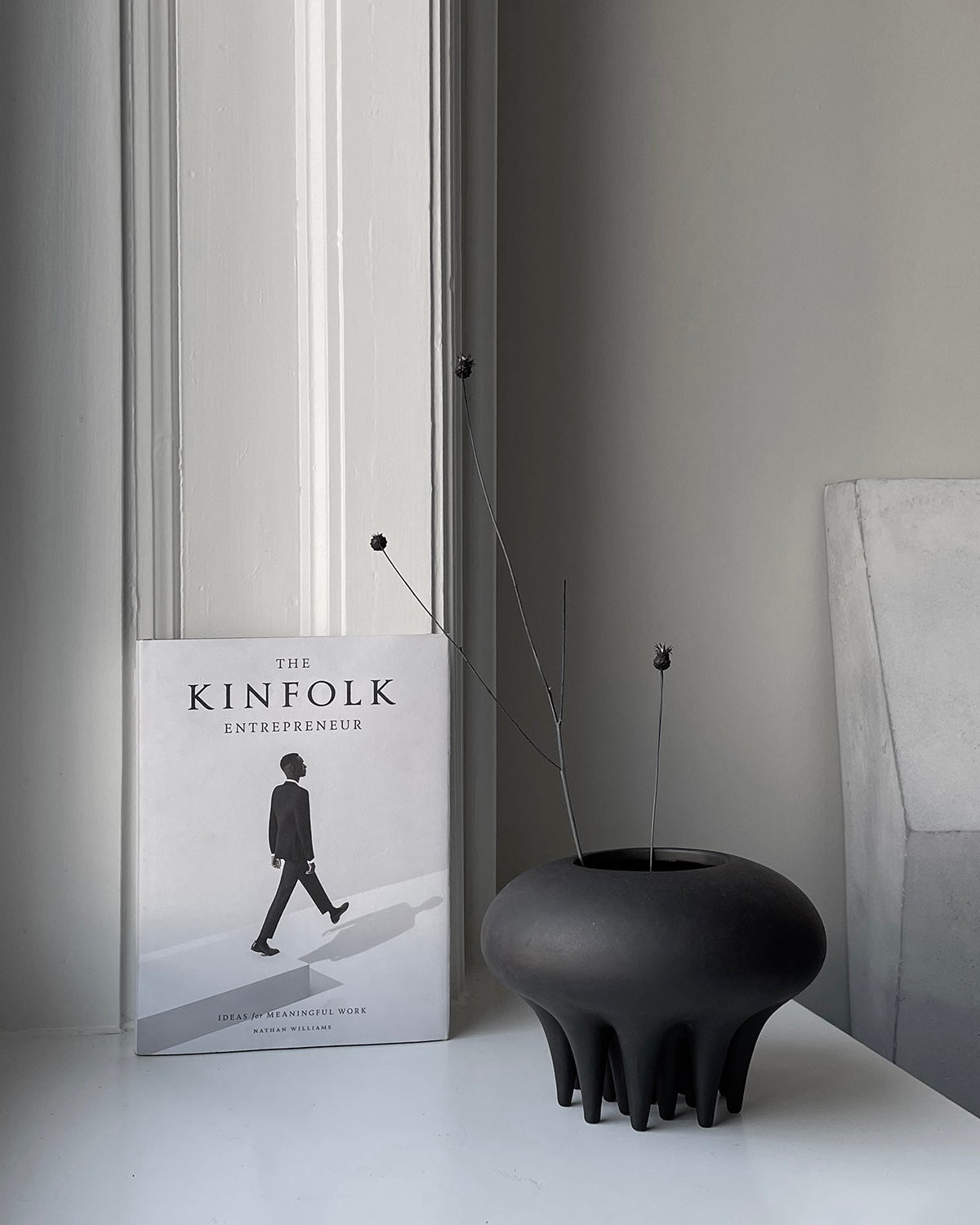 Book titled 'The Kinfolk Entrepreneur' on a surface with a black vase. Medusa Vase, Mini by 101 Copenhagen. Sculptural ceramic mini vase in Coffee with soft oval shapes and conical legs, hand-glazed and not 100% waterproof – curated by Numa Home.