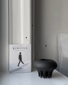 Book titled 'The Kinfolk Entrepreneur' on a surface with a black vase. Medusa Vase, Mini by 101 Copenhagen. Sculptural ceramic mini vase in Coffee with soft oval shapes and conical legs, hand-glazed and not 100% waterproof – curated by Numa Home.