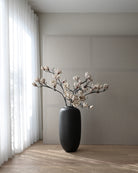 Black vase with white lily flowers on a wooden floor against a gray wall. Sunao Vase Slim, Hexa by 101 Copenhagen, curated by Numa Home