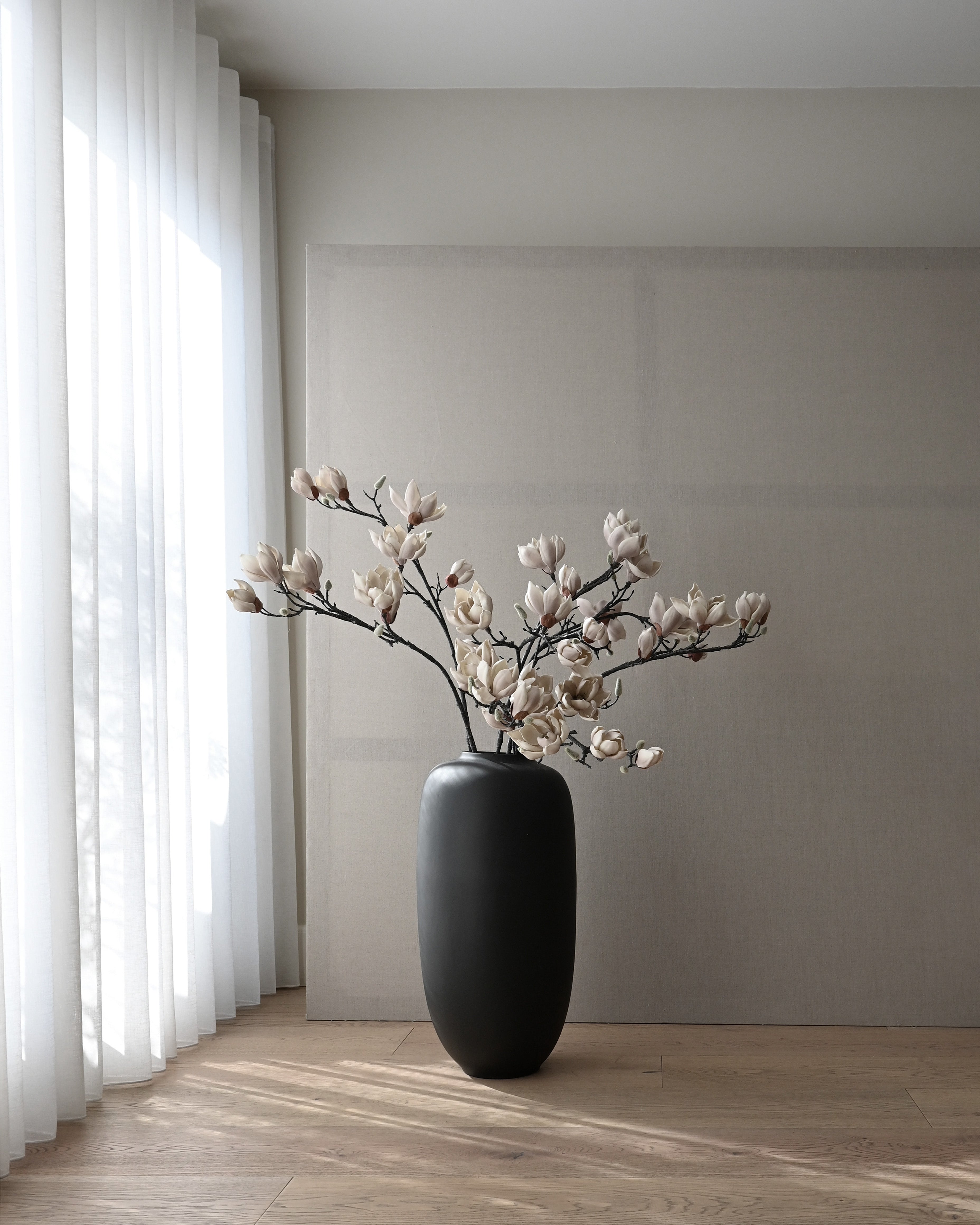 Black vase with white lily flowers on a wooden floor against a gray wall. Sunao Vase Slim, Hexa by 101 Copenhagen, curated by Numa Home