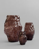 Three brown ceramic vases of different sizes on a gray background. Katachi Vase, Big by 101 Copenhagen. Sculptural vase inspired by Japanese “shape”, available in High Glaze Birch ceramic and Clear or Smoked Forest mouth-blown glass. Fully waterproof, curated by Numa Home.