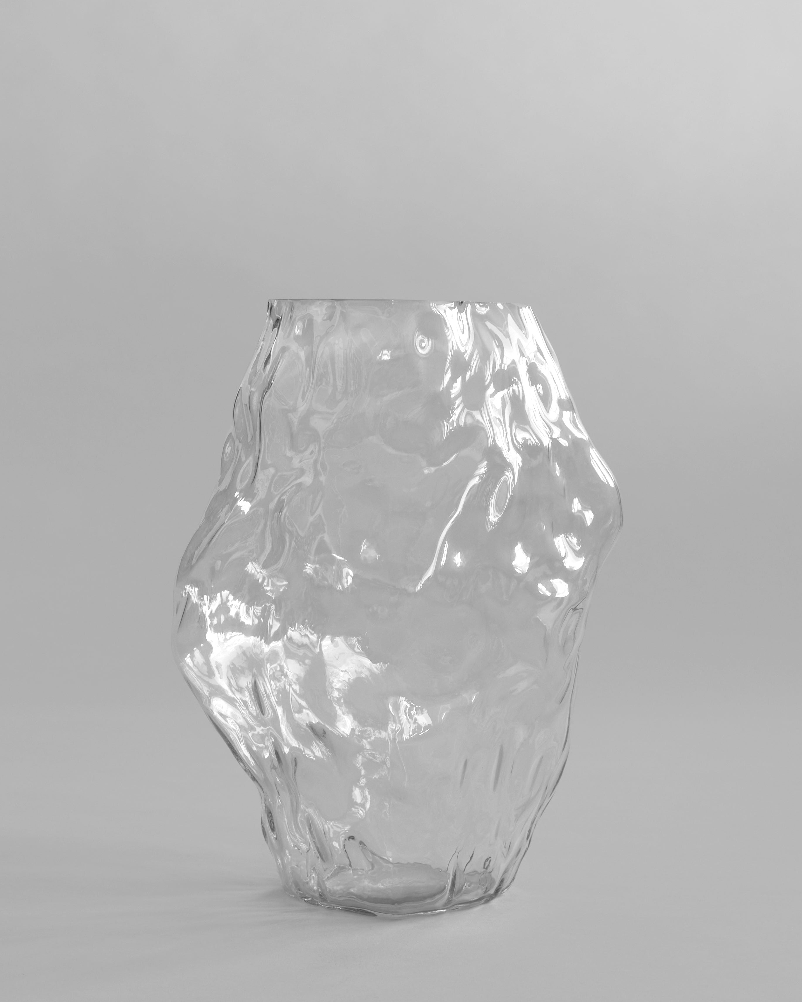 Clear glass vase on a gray background. Katachi Vase, Big by 101 Copenhagen. Sculptural vase inspired by Japanese “shape”, available in High Glaze Birch ceramic and Clear or Smoked Forest mouth-blown glass. Fully waterproof, curated by Numa Home.