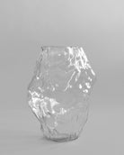 Clear glass vase on a gray background. Katachi Vase, Big by 101 Copenhagen. Sculptural vase inspired by Japanese “shape”, available in High Glaze Birch ceramic and Clear or Smoked Forest mouth-blown glass. Fully waterproof, curated by Numa Home.