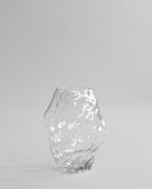 Clear glass vase on a light gray background. Katachi Vase, Medio by 101 Copenhagen. Sculptural vase inspired by Japanese “shape”, available in High Glaze Birch ceramic and Clear or Smoked Forest mouth-blown glass. Fully waterproof – curated by Numa Home.