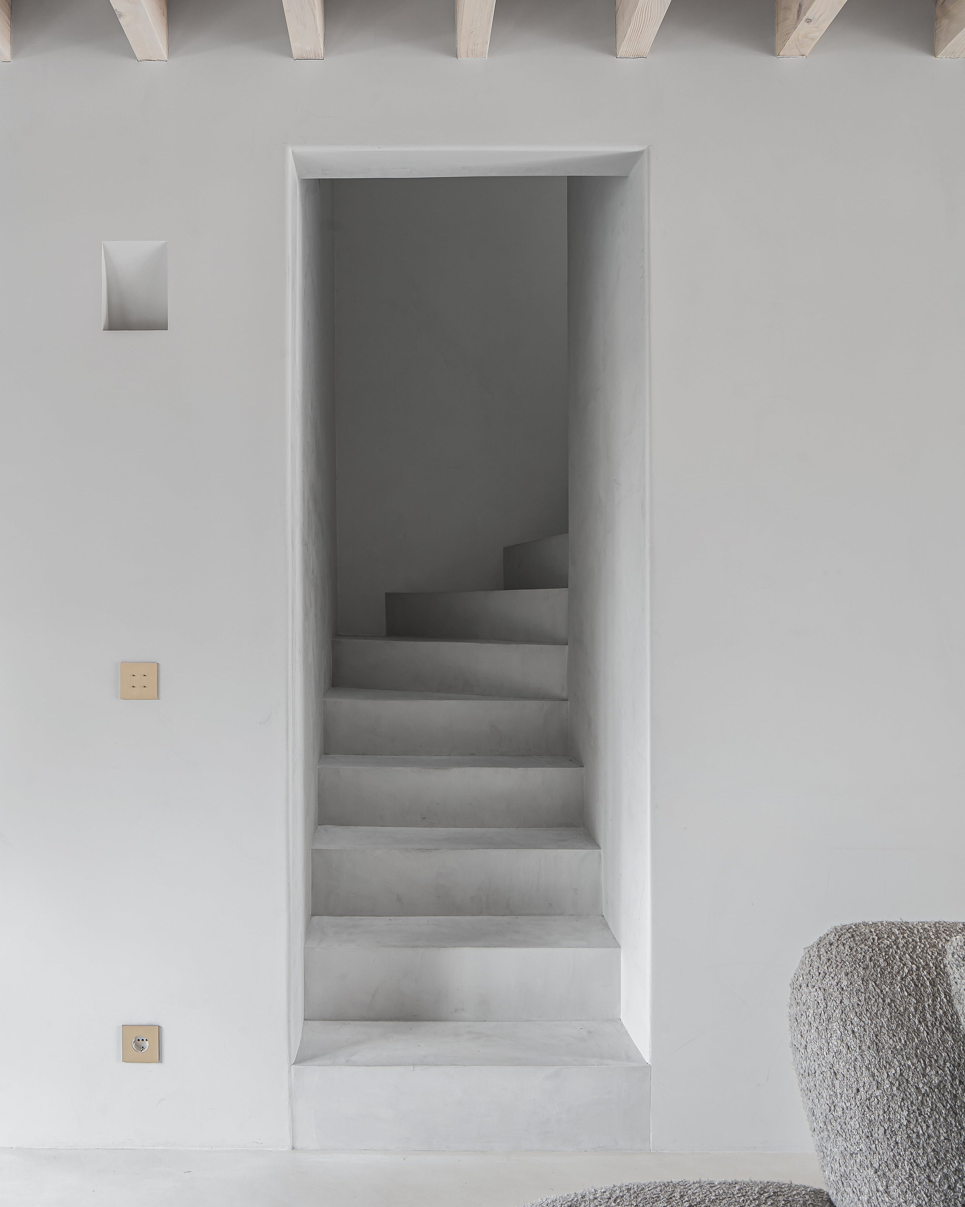 Stairwell opening in a white wall with wooden beams on the ceiling. Sumum PIN switch with four buttons on a single plate in brushed brass color
