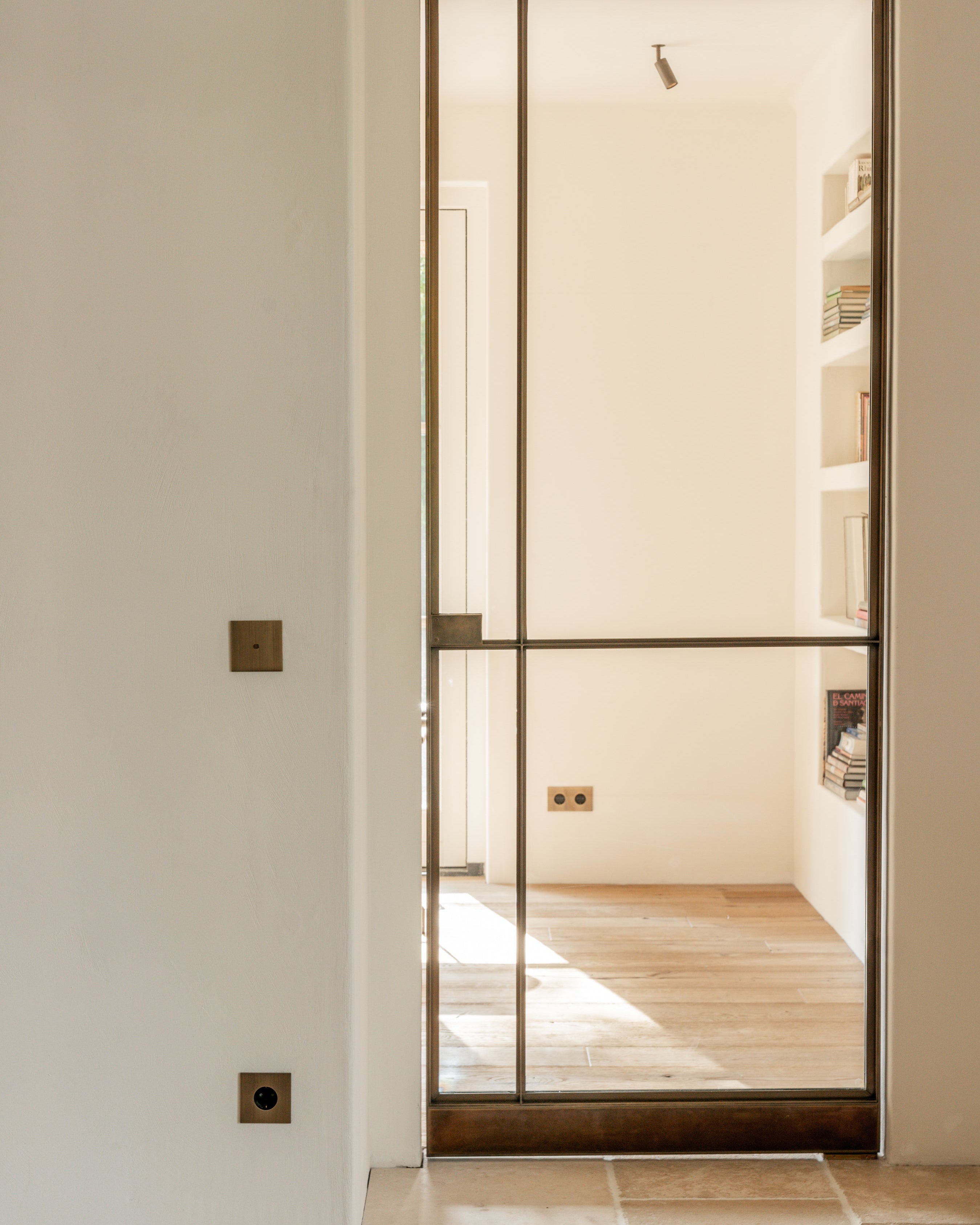 Modern glass door with a metal frame in a neutral interior setting. Brushed anique bronze switches, outlets by Sumum, and a brass LED spotlight in wonderful harmony as the same finish.
