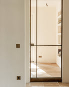 Modern glass door with a metal frame in a neutral interior setting. Brushed anique bronze switches, outlets by Sumum, and a brass LED spotlight in wonderful harmony as the same finish.