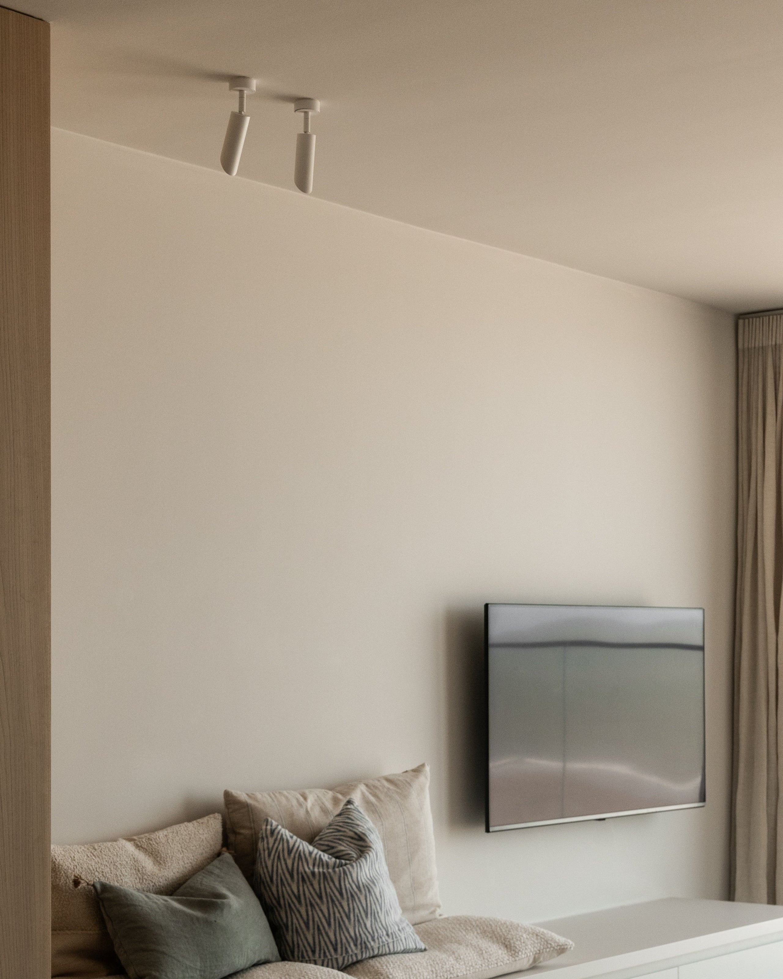 Modern living room with a television mounted on the wall, a sofa with pillows, and a window with curtains. White brass ceiling light fixture with bevelled nose. Sumum brass spotlights made from brass partnered with PSM Lighting