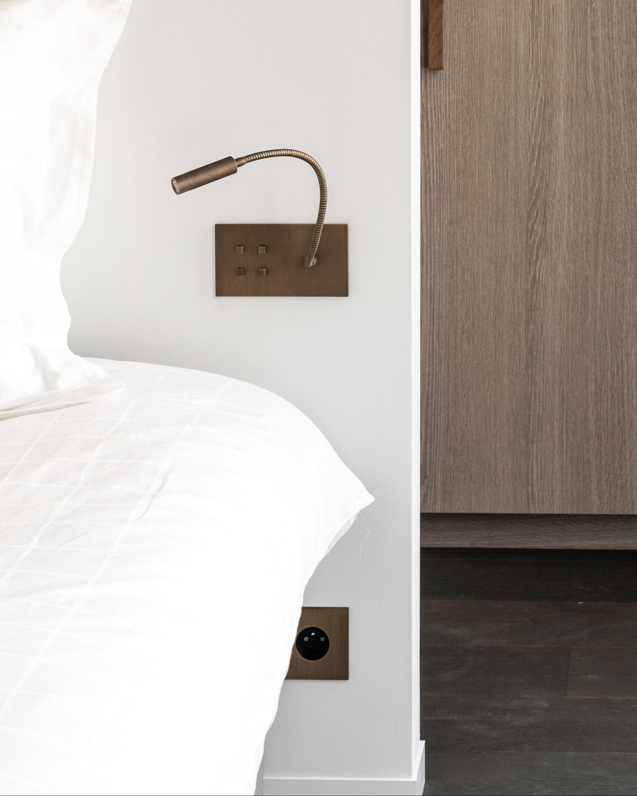 Bedroom with wooden wardrobe and white bedding. Brass reading light in brushed bronze patina with 4 buttons to control the lights.