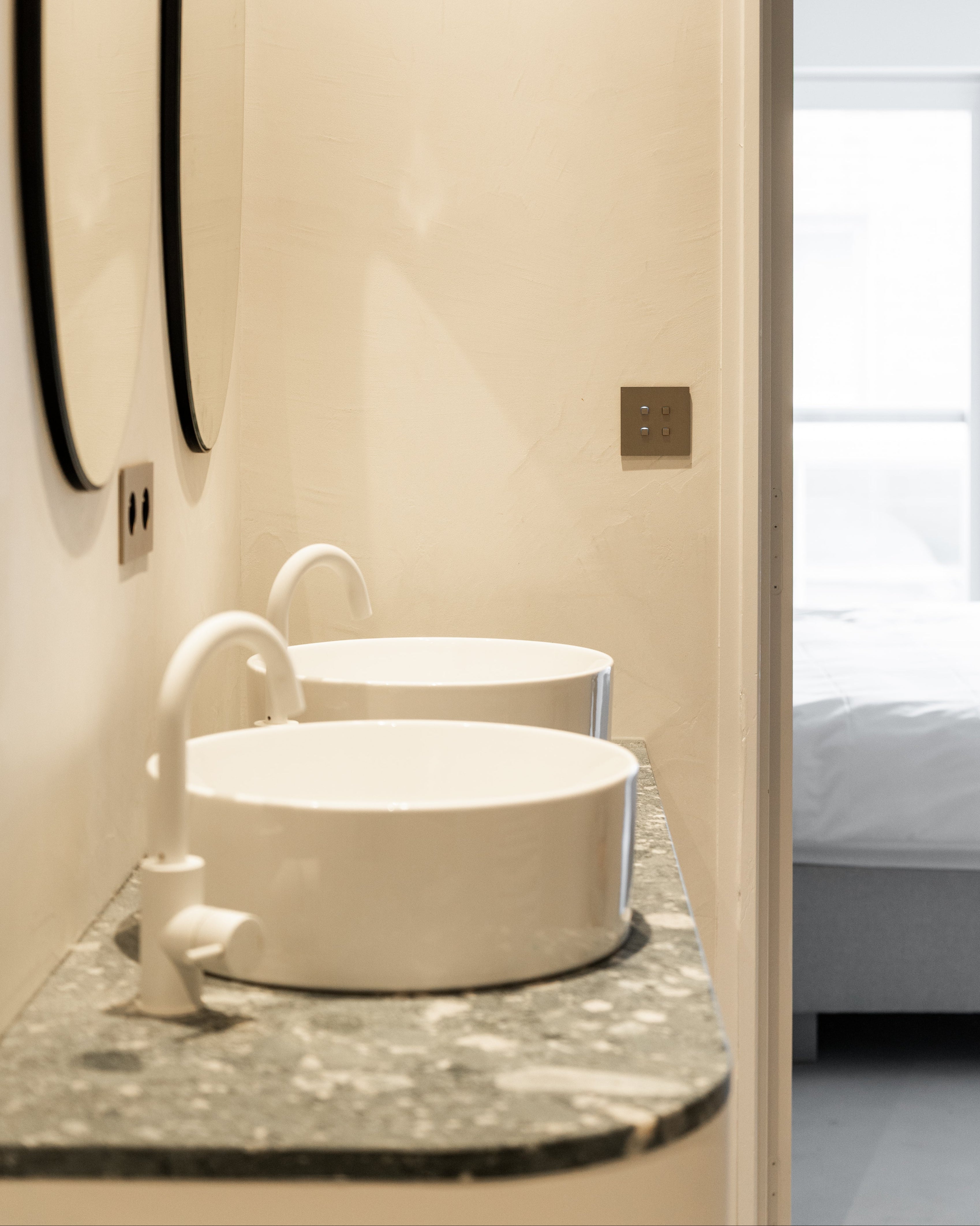 Sumum Square brushed brass switch mounted in a modern bathroom with marble countertop and twin white basins, premium custom light switch for luxury interiors provided by Numa Home