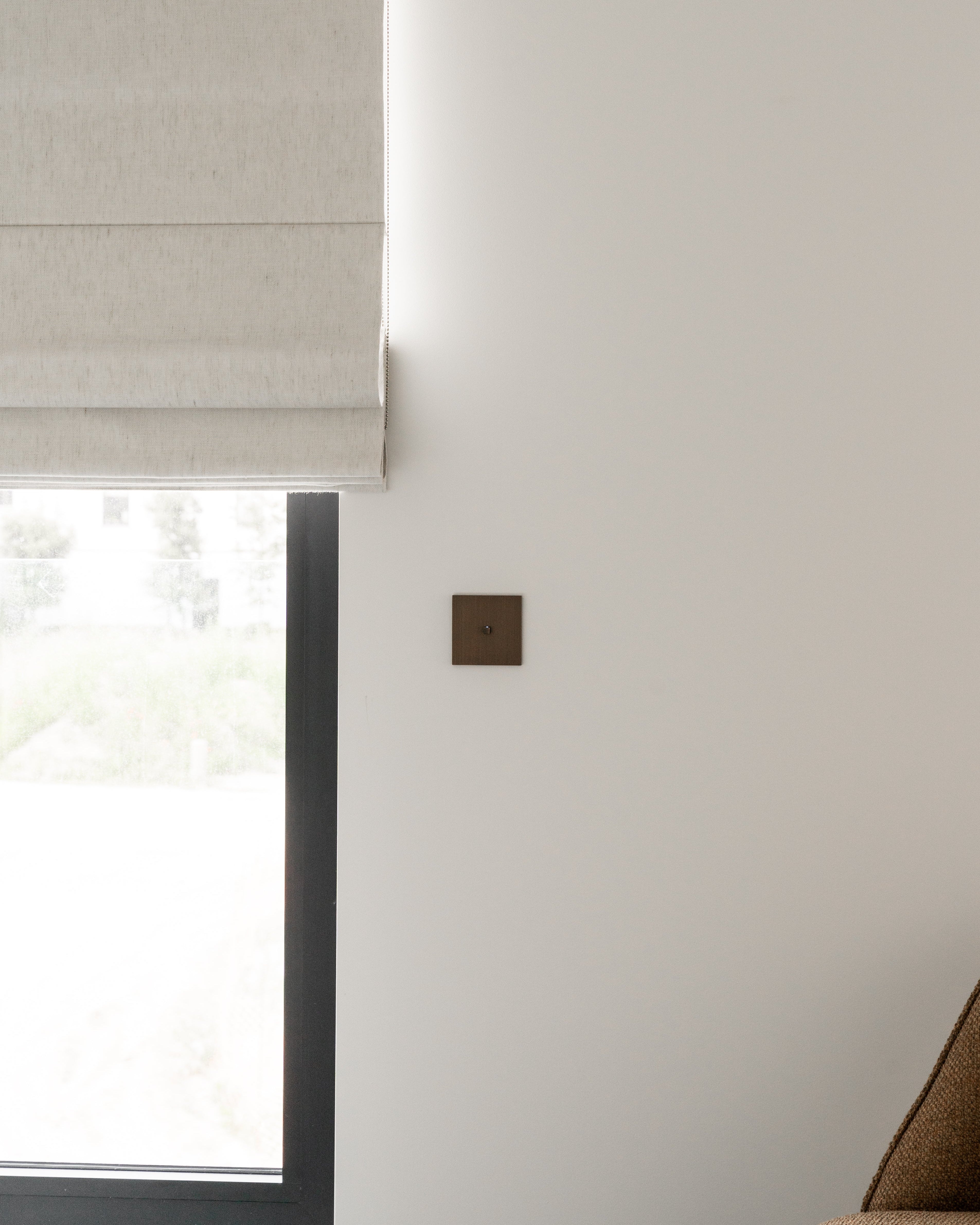Sumum Square single-button brass switch in dark bronze finish, mounted beside a window with a roman shade in a modern Dutch interior, custom high-end light switch provided by Numa Home